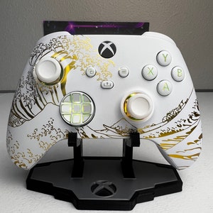 May include: White Xbox controller with gold accents and a wave design. The controller is on a white stand with a black Xbox logo.