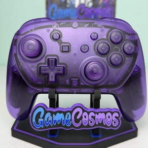 May include: A translucent purple wireless video game controller featuring two analog sticks, a directional pad, and action buttons. The controller rests on a black stand with a blue and purple sign that reads GameCosmos.