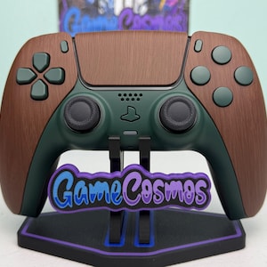 May include: A wireless video game controller featuring a dark green base with brown wood-grain textured side panels and touchpad. The controller is mounted on a black stand with purple accents and a GameCosmos logo plate attached to the front.