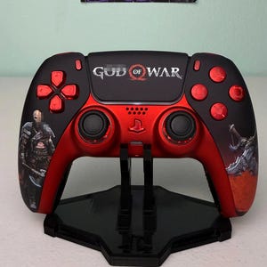 May include: A custom red and black video game controller featuring God of War branding and character graphics. The controller has metallic red buttons, a central touchpad with the game logo, and black analog sticks, mounted on a black stand.