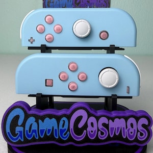 Könnte beinhalten: Zwei hellblaue Gamecontroller mit rosa Knöpfen und weißen Joysticks sind auf einem schwarzen Ständer gestapelt. Der Ständer hat ein lila-blaues "Game Cosmos"-Logo. Die Controller sind für Spiele konzipiert.