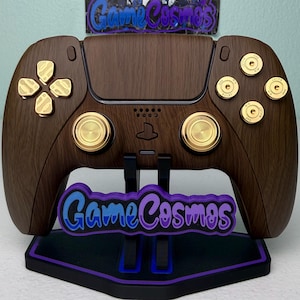 May include: A wireless video game controller with a dark brown wood grain finish, featuring gold-colored metallic buttons, directional pad, and analog sticks. The controller is mounted on a black stand with a purple GameCosmos logo plate.