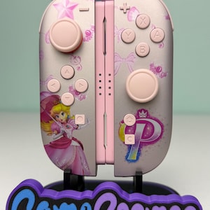 May include: Pink and silver Nintendo Switch Joy-Cons with a Princess Peach design. The controllers have pink buttons and a floral pattern. The base has the text "Game Cosmos" in blue and purple.