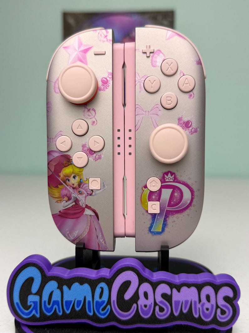 May include: Pink and silver Nintendo Switch Joy-Cons with a Princess Peach design. The controllers have pink buttons and a floral pattern. The base has the text "Game Cosmos" in blue and purple.
