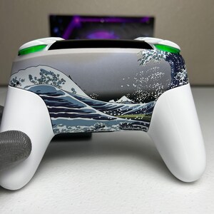 Puede incluir: Un mando de videojuegos blanco con un dise&ntilde;o personalizado que presenta un patr&oacute;n de olas azules y blancas. El mando tiene botones verdes en la parte superior.