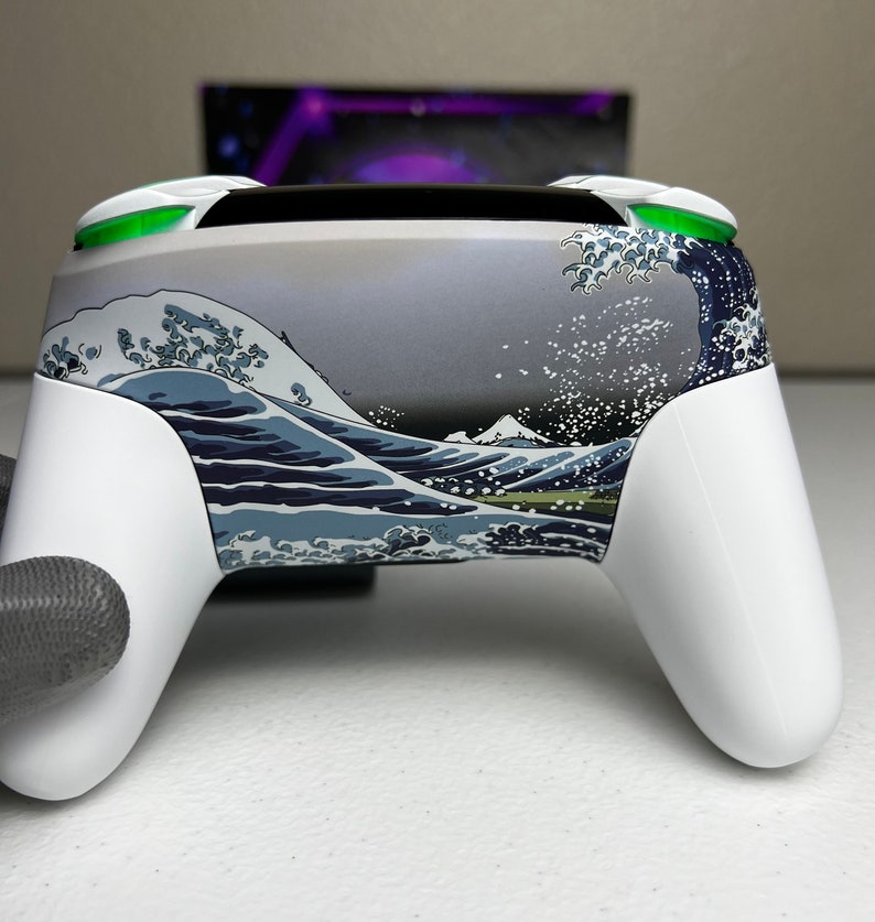 Puede incluir: Un mando de videojuegos blanco con un dise&ntilde;o personalizado que presenta un patr&oacute;n de olas azules y blancas. El mando tiene botones verdes en la parte superior.