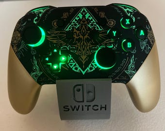 May include: A black and gold wireless video game controller featuring glowing green LED accents, a central intricate gold graphic design, and illuminated A, B, X, and Y buttons. The controller rests on a grey stand embossed with the word SWITCH.