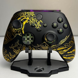 Op de afbeelding: Een zwarte en gouden Xbox-controller met een golfontwerp. De controller staat op een zwarte standaard met een grijs Xbox-logo.