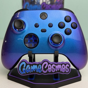 May include: A wireless video game controller with a metallic blue and purple gradient finish. The controller features black analogue sticks, a black directional pad, and black action buttons labelled X, Y, A, and B. It rests on a black stand with the text GameCosmos.