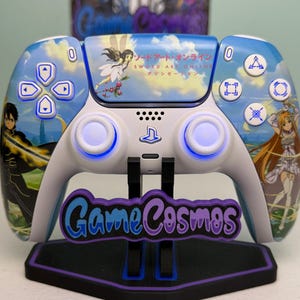 May include: A white wireless game controller featuring Sword Art Online graphics, including anime characters and a fairy on the touchpad. The controller has blue light-up analog sticks, directional buttons, and action buttons. It is displayed on a black stand with the purple and blue text GameCosmos.