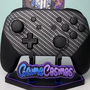 Puede incluir: Un mando de videojuegos con un diseño de fibra de carbono, botones negros y una cruceta. El mando está sobre un soporte con el texto "Game Cosmos" en azul y morado.