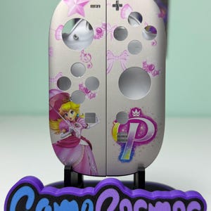 May include: Custom-designed Nintendo Switch Joy-Con controllers featuring a Princess Peach theme. The controllers are light gray with pink accents, including stars, bows, and a graphic of Princess Peach. The base features the text "Game Cosmos" in purple and blue.