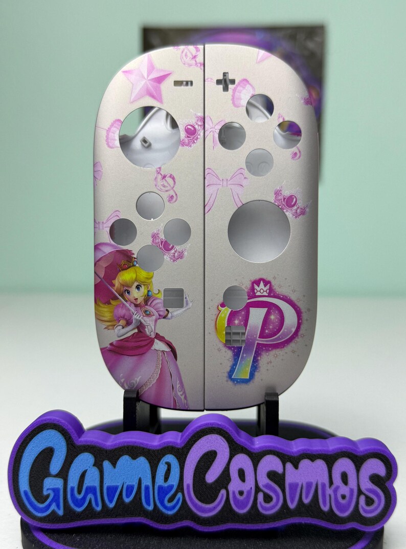 May include: Custom-designed Nintendo Switch Joy-Con controllers featuring a Princess Peach theme. The controllers are light gray with pink accents, including stars, bows, and a graphic of Princess Peach. The base features the text "Game Cosmos" in purple and blue.