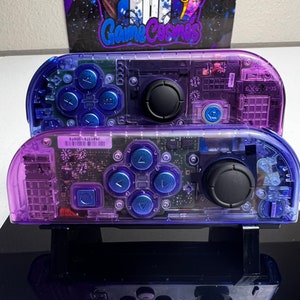 May include: Two clear purple Nintendo Switch controllers with blue buttons and black joysticks. The controllers are translucent and show the internal circuitry. The top controller has the text "39878-100876" printed on it.