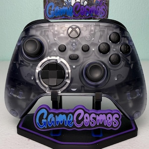 Peut inclure: Une manette Xbox transparente avec des boutons noirs et un pavé directionnel noir, posée sur un support. Le support est noir avec des accents violets et bleus et le texte "GameCosmos". Les composants internes de la manette sont visibles.