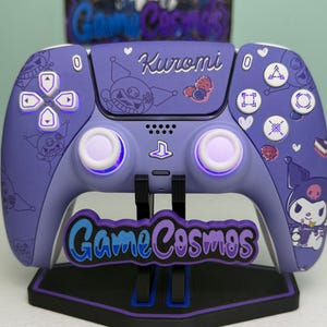 Op de afbeelding: Een paarse gamecontroller met bijpassende standaard. De controller heeft witte knoppen en een ontwerp met het woord "Kuromi" en stripfiguren. De standaard heeft de woorden "Game Cosmos" in een blauw en paars ontwerp.