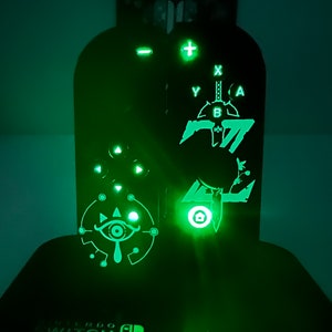 Może przedstawiać: Czarna stacja dokująca Nintendo Switch z zielonym podświetleniem. Stacja dokująca ma wzór przedstawiający Triforce i Master Sword z serii gier wideo The Legend of Zelda. Stacja dokująca ma również zielone światło z przodu.