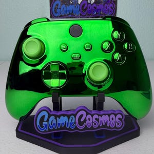 Könnte beinhalten: Ein glänzend grüner Xbox-Controller mit passenden grünen Knöpfen und Thumbsticks. Der Controller steht auf einem schwarzen Ständer mit dem Text "Game Cosmos" in Lila und Blau. Der Controller hat ein metallisches Finish.