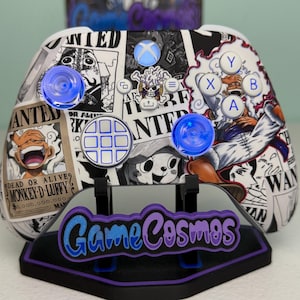 May include: A custom Xbox wireless controller featuring a black and white collage of One Piece wanted posters, including Monkey D. Luffy. The controller has translucent blue thumbsticks, white action buttons, and a circular directional pad.