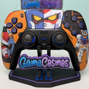 May include: A black PlayStation 5 controller with a custom design featuring robot illustrations in orange, blue, and white. The controller is on a stand with the text "Game Cosmos".