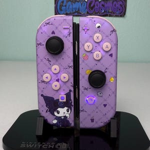 Könnte beinhalten: Ein Paar lavendelfarbene Nintendo Switch Joy-Con-Controller mit einem durchgehenden Rautenmuster, Schleifen, Herzen und kleinen Lebensmittelsymbolen. Der linke Controller zeigt die Figur Kuromi, beide haben hellrosa Tasten.