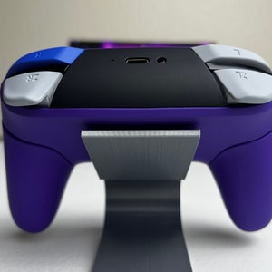 May include: A purple video game controller with white buttons and a black top is sitting on a gray stand.