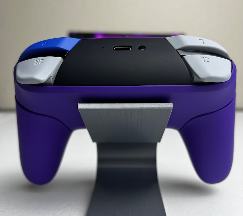 May include: A purple video game controller with white buttons and a black top is sitting on a gray stand.