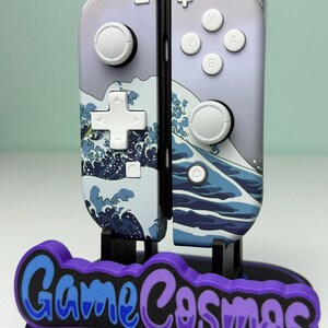 May include: A pair of Nintendo Switch Joy-Con controllers with a custom design featuring a wave pattern in shades of blue and white. The controllers are displayed on a black stand with the text "Game Cosmos" in blue and purple.