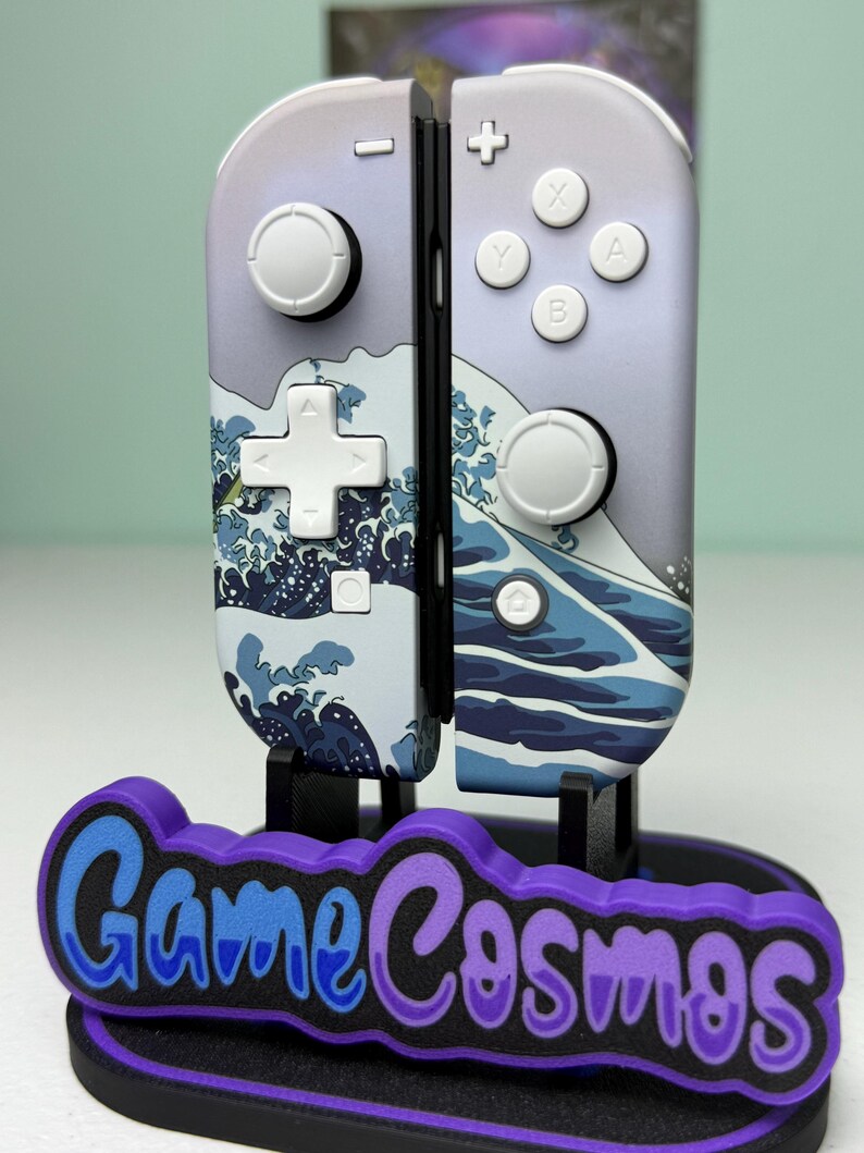 May include: A pair of Nintendo Switch Joy-Con controllers with a custom design featuring a wave pattern in shades of blue and white. The controllers are displayed on a black stand with the text "Game Cosmos" in blue and purple.