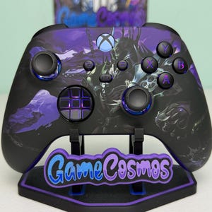 Op de afbeelding: Zwarte en paarse Xbox-controller met een fantasie-ontwerp. De controller heeft een gedetailleerde afbeelding en wordt weergegeven op een standaard met de tekst "Game Cosmos". De knoppen zijn gekleurd in paars, blauw en groen.