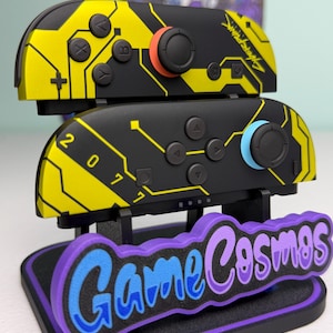 Puede incluir: Mandos Joy-Con de Nintendo Switch negros y amarillos con tem&aacute;tica Cyberpunk, con detalles en naranja y azul. Los mandos se muestran en un soporte con el texto "Game Cosmos" en azul y morado.