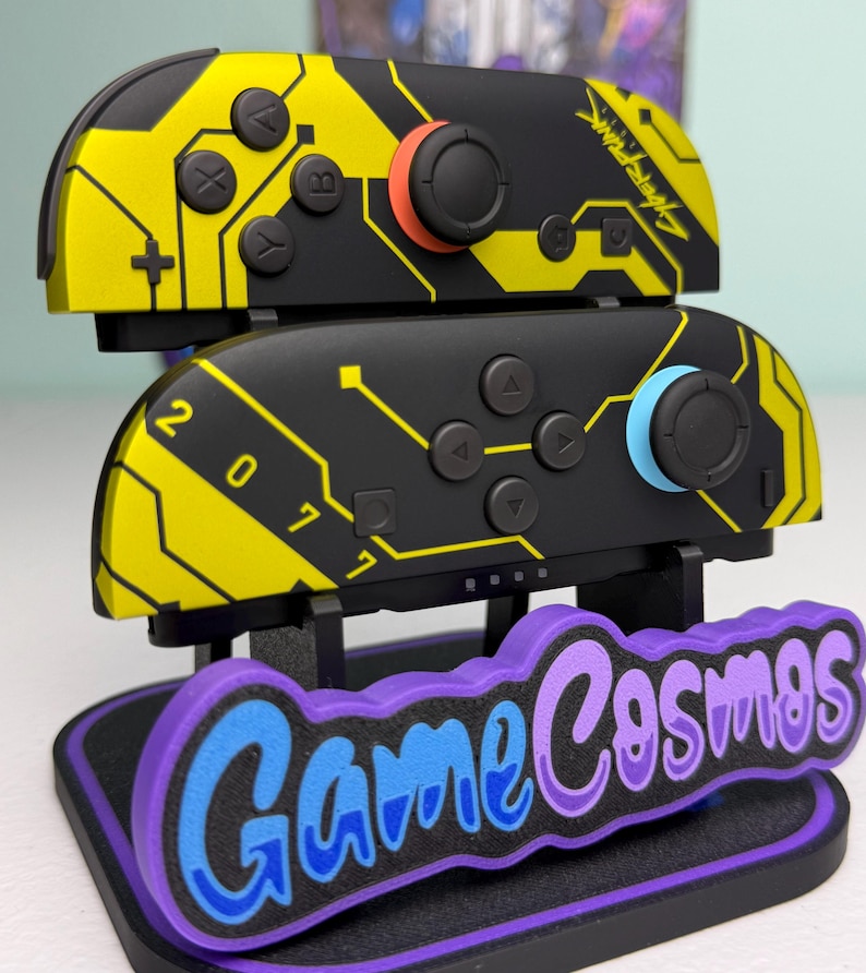 Puede incluir: Mandos Joy-Con de Nintendo Switch negros y amarillos con tem&aacute;tica Cyberpunk, con detalles en naranja y azul. Los mandos se muestran en un soporte con el texto "Game Cosmos" en azul y morado.