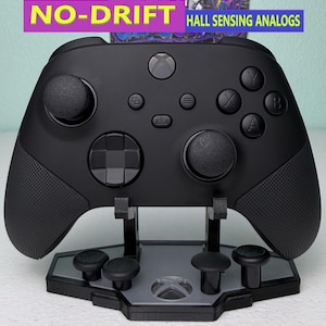 Może przedstawiać: Czarny kontroler Xbox z analogowymi gałkami z czujnikami Halla, na czarnym stojaku z szarym logo Xbox. Kontroler ma teksturowany uchwyt i jest w nowym stanie.