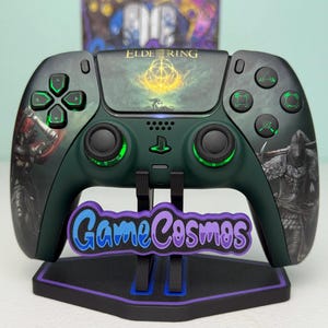 May include: A dark green video game controller featuring Elden Ring graphics, including a central golden emblem and character illustrations on the grips. The controller has glowing green accents on the buttons and joysticks, and it is mounted on a black stand with a purple and blue GameCosmos logo.