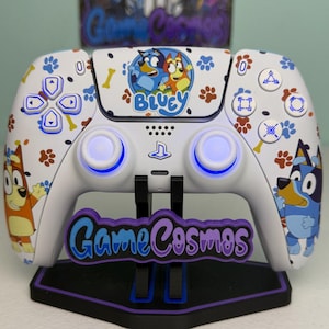 May include: A white video game controller featuring Bluey character graphics, paw prints, and bone patterns. The center touchpad displays the Bluey logo. The controller is mounted on a black stand with a purple GameCosmos logo plate attached.