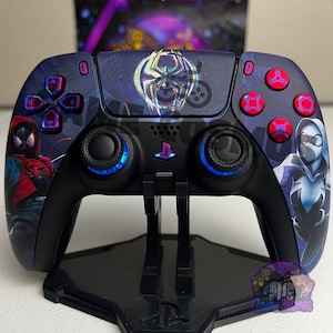 May include: A black PlayStation 5 controller with a custom design featuring Spider-Man characters. The controller has blue and red accents and is displayed on a black stand.