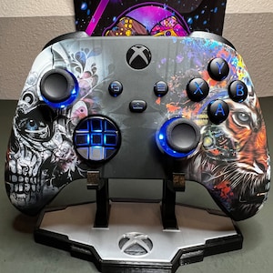 Könnte beinhalten: Ein schwarz-weißer Xbox-Controller mit Totenkopf- und Tigerdesign. Der Controller hat blaue Knöpfe und steht auf einem Ständer.