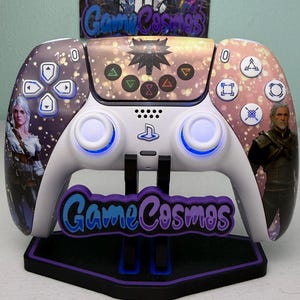 Pode incluir: Um controle PlayStation 5 branco e roxo com um design personalizado com personagens e símbolos da série de jogos The Witcher. O controle tem um suporte correspondente com o texto "Game Cosmos".