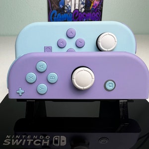 Może przedstawiać: Dwa kontrolery Joy-Con do Nintendo Switch, jeden jasnoniebieski, a drugi lawendowy, z logo Nintendo Switch na dole. Kontrolery są ułożone jeden na drugim.