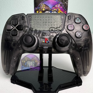 Puede incluir: Un mando PlayStation 5 DualSense translúcido negro con un patrón de cuadros en el panel táctil. El mando está en un soporte negro con el logotipo de PlayStation.