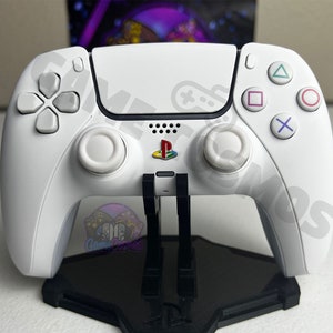 May include: A white PlayStation 5 DualSense controller is displayed on a black stand. The controller has a white faceplate with the PlayStation logo and a black faceplate with the buttons. The controller is in a vertical position with the buttons facing the viewer.