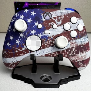 May include: An Xbox controller with a red, white and blue American flag design. The controller has a distressed look with white buttons and a white D-pad. It is displayed on a stand.
