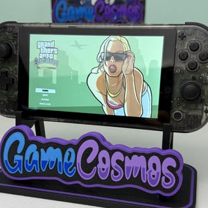 以下が含まれることがあります： Grand Theft Auto: San Andreasのゲームを表示する透明な携帯ゲーム機。デバイスは、紫と青の「Game Cosmos」ロゴが付いた黒いスタンドに取り付けられています。ゲーム画面にはサングラスをかけた女性が表示されています。