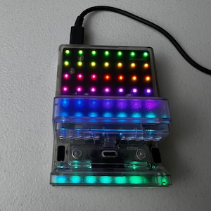 Può includere: Un dispositivo elettronico rettangolare trasparente con un cavo nero collegato. Il dispositivo ha più file di luci LED colorate, che mostrano uno spettro di colori. Il dispositivo è su una superficie bianca.