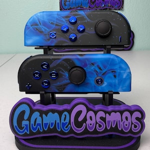 May include: A black and blue gaming controller set with a stand. The controllers have a blue smoke design and blue buttons. The stand has the text "Game Cosmos" in blue and purple.