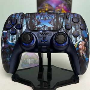 Puede incluir: Un mando de PlayStation 5 azul y negro con un diseño personalizado que presenta a Deadpool y Wolverine. El mando tiene un fondo de bosque azul y negro con las palabras "Weapon X" en blanco. El mando está en un soporte negro.