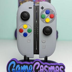 May include: A pair of light gray video game controllers featuring a retro-inspired layout with multicolored action buttons, black joysticks, and directional inputs. The controllers are mounted on a black stand with the text GameCosmos in purple.