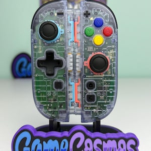 Può includere: Un controller per videogiochi trasparente con componenti interni visibili, joystick neri e pulsanti colorati. Il controller è montato su un supporto con il testo "Game Cosmos" in viola e blu.