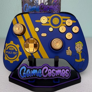 May include: A blue and yellow wireless game controller featuring a Vault-Tec gear logo, a Vault Boy character giving a thumbs up, and gold-colored buttons shaped like bullet casings. The controller is displayed on a black stand with a GameCosmos logo.