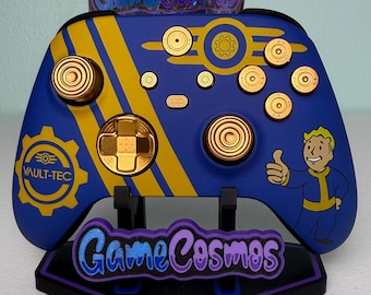 Puede incluir: Un mando de juego inalámbrico azul y amarillo con el logotipo de engranaje de Vault-Tec, un personaje Vault Boy haciendo un gesto de pulgar hacia arriba y botones dorados con forma de casquillos de bala sobre un soporte negro con el logo GameCosmos.